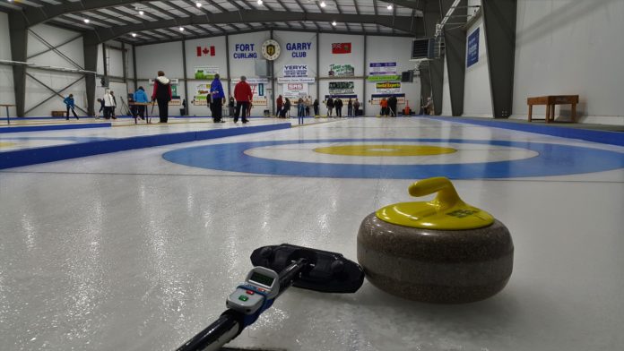 A curling rock sits next to a curling brush on the ice, waiting to be thrown down to the waiting house.