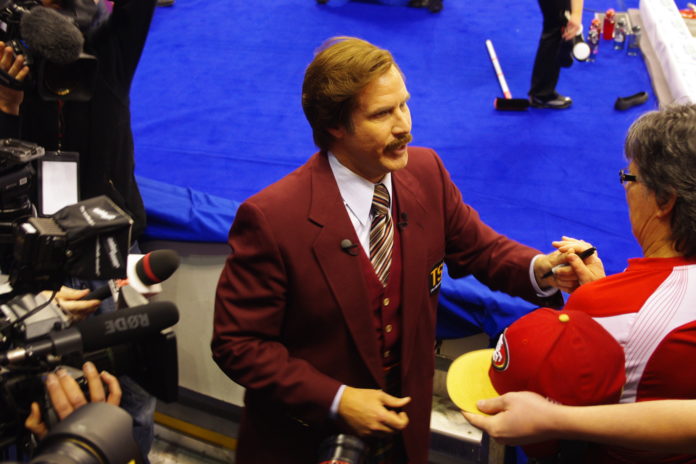 Fictional news anchorman Ron Burgundy, played by Will Ferrell, is wearing a burgundy suit jacket and signing autographs for fans at the Olympic curling trials in Winnipeg.