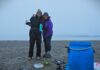 Wilderness adventure comes full circle for aunty and niece Two women in outdoor clothing shells - one wearing blue, one wearing purple - standing together near a foggy shoreline. Older woman (50-ish) has arm around younger woman (20-ish). They just finished a breakfast of oatmeal and coffee, and are about to pack up their big blue food barrel, also seen in the frame.
