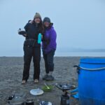 Wilderness adventure comes full circle for aunty and niece Two women in outdoor clothing shells - one wearing blue, one wearing purple - standing together near a foggy shoreline. Older woman (50-ish) has arm around younger woman (20-ish). They just finished a breakfast of oatmeal and coffee, and are about to pack up their big blue food barrel, also seen in the frame.