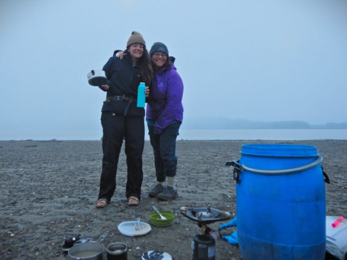 Two women in outdoor clothing shells - one wearing blue, one wearing purple - standing together near a foggy shoreline. Older woman (50-ish) has arm around younger woman (20-ish). They just finished a breakfast of oatmeal and coffee, and are about to pack up their big blue food barrel, also seen in the frame.