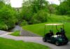 Minnesota golf courses perfect for self-guided golf tours A dark green golf cart is present in the lower right corner of the photo, parked beside an elevated tee box, overlooking a cart trail that slopes downward to a bridge that covers a water hazard. Just one of over 400 Minnesota golf courses in the state.