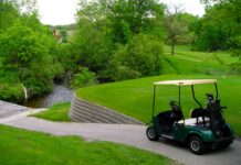 Minnesota golf courses perfect for self-guided golf tours A dark green golf cart is present in the lower right corner of the photo, parked beside an elevated tee box, overlooking a cart trail that slopes downward to a bridge that covers a water hazard. Just one of over 400 Minnesota golf courses in the state.