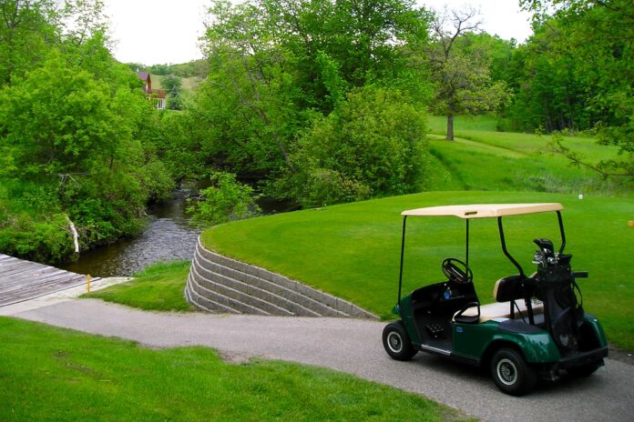 A dark green golf cart is present in the lower right corner of the photo, parked beside an elevated tee box, overlooking a cart trail that slopes downward to a bridge that covers a water hazard. Just one of over 400 Minnesota golf courses in the state.