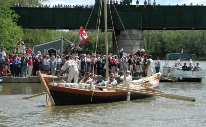 A large brown york boat with a mast and six rowers pushes away from a public dock where hundreds of people gather to watch the start of this unique and bizarre york boat voyage.