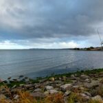 Beach Roaming: Dublin, Ireland The view of a rock and mossy beach stretching along the blue-grey ocean, with dark clouds above, and a thin band of blue sky off on the horizon. It's one of a half dozen Dublin city beaches.
