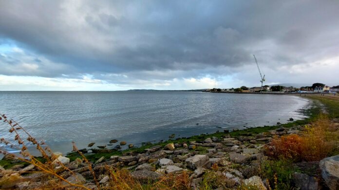 The view of a rock and mossy beach stretching along the blue-grey ocean, with dark clouds above, and a thin band of blue sky off on the horizon. It's one of a half dozen Dublin city beaches.