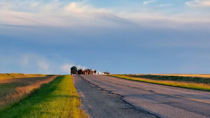 A film crew making The Long Walk, on the horizon in motorized carts at golden hour, in a prairie location, on an old concrete highway, with green and yellow fields, and a deep blue sunset sky.