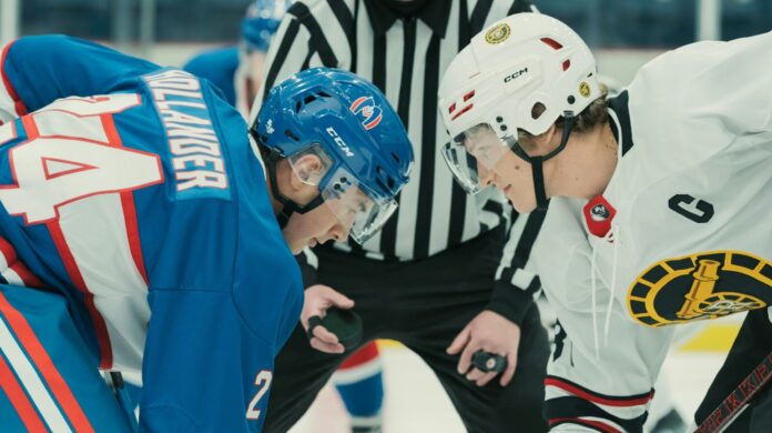 Two hockey players facing off the puck, each one crouched down low, and face to face. One player looks down at the puck. The other gazes at him.