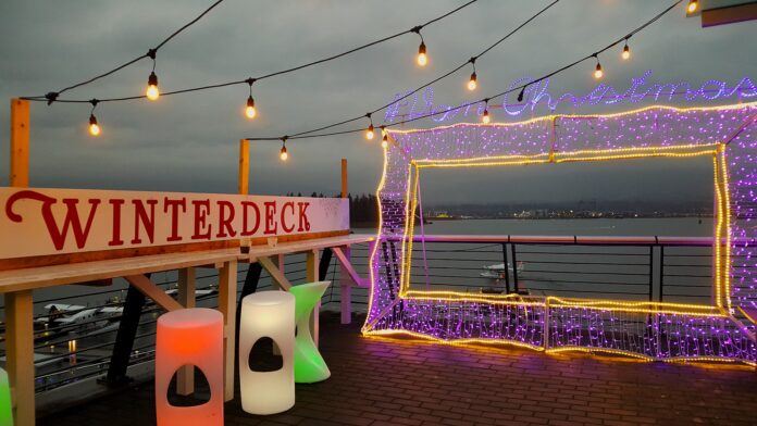 A picture frame made from purple LED lights is on a deck, with the sign'winterdeck' beside it, and an ocean view behind it.