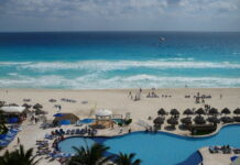 Winter vacations in Mexico along the Yucatan Peninsula White rolling waves on a turquoise colored ocean, and white sandy beach, with a swimming pool and pool chairs in the foreground.