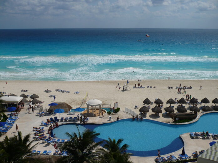 White rolling waves on a turquoise colored ocean, and white sandy beach, with a swimming pool and pool chairs in the foreground.