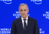 Mark Carney addresses the world from Davos, Switzerland Prime Minister Mark Carney in a dark blue suit and tie, white shirt, and order of Canada pin on his lapel, addresses the audience from a podium on a stage, in front of a royal blue backdrop with 'World Economic Forum' branding on it.