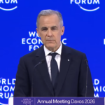 Mark Carney addresses the world from Davos, Switzerland Prime Minister Mark Carney in a dark blue suit and tie, white shirt, and order of Canada pin on his lapel, addresses the audience from a podium on a stage, in front of a royal blue backdrop with 'World Economic Forum' branding on it.