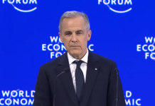 Mark Carney addresses the world from Davos, Switzerland Prime Minister Mark Carney in a dark blue suit and tie, white shirt, and order of Canada pin on his lapel, addresses the audience from a podium on a stage, in front of a royal blue backdrop with 'World Economic Forum' branding on it.