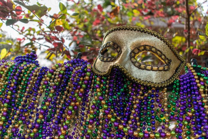 A golden mardi gras face mask sits atop a fence draped with colourful beads.
