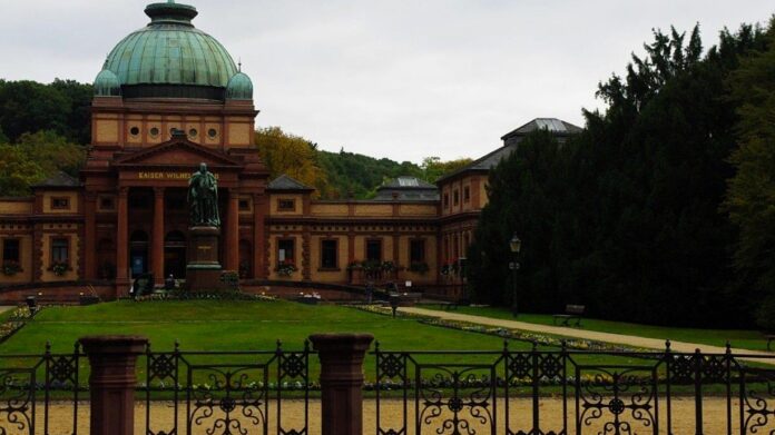The stone-clad exterior of a spa facility in the German town of Bad Homburg, nestled inside a forested park in the centre of the city, behind iron fencing and rows of flowers and foliage.