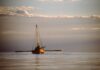 Quest for the Bay: 25 years since the big adventure A wooden boat with a mast and outstretched oars glides across a flat lake with picturesque clouds in the sky.