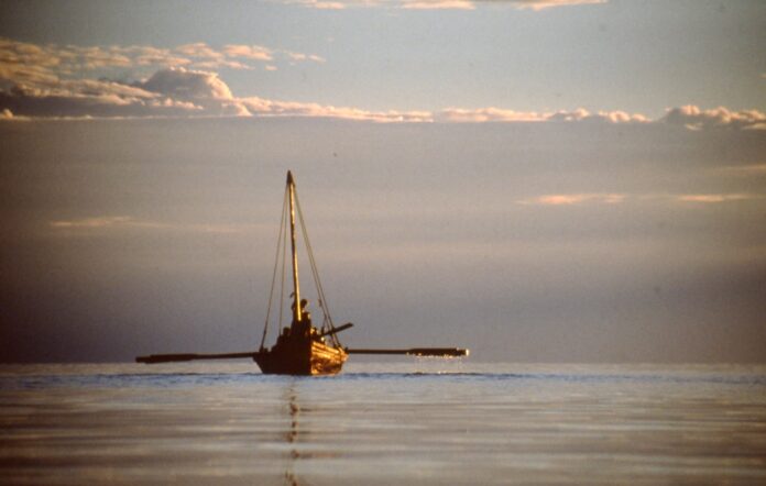 A wooden boat with a mast and outstretched oars glides across a flat lake with picturesque clouds in the sky.