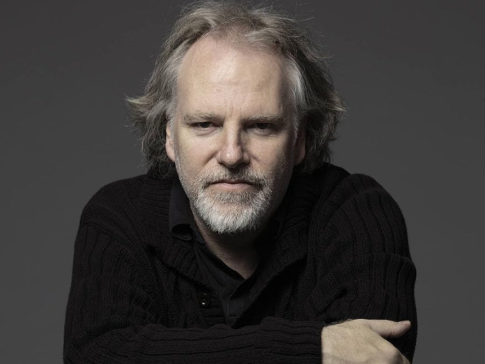 Close-up shot of Guy Maddin, a distinguished looking man with thinning grey hair and goatee, wearing a black sweater, arms crossed in front, leaning into and looking into camera.